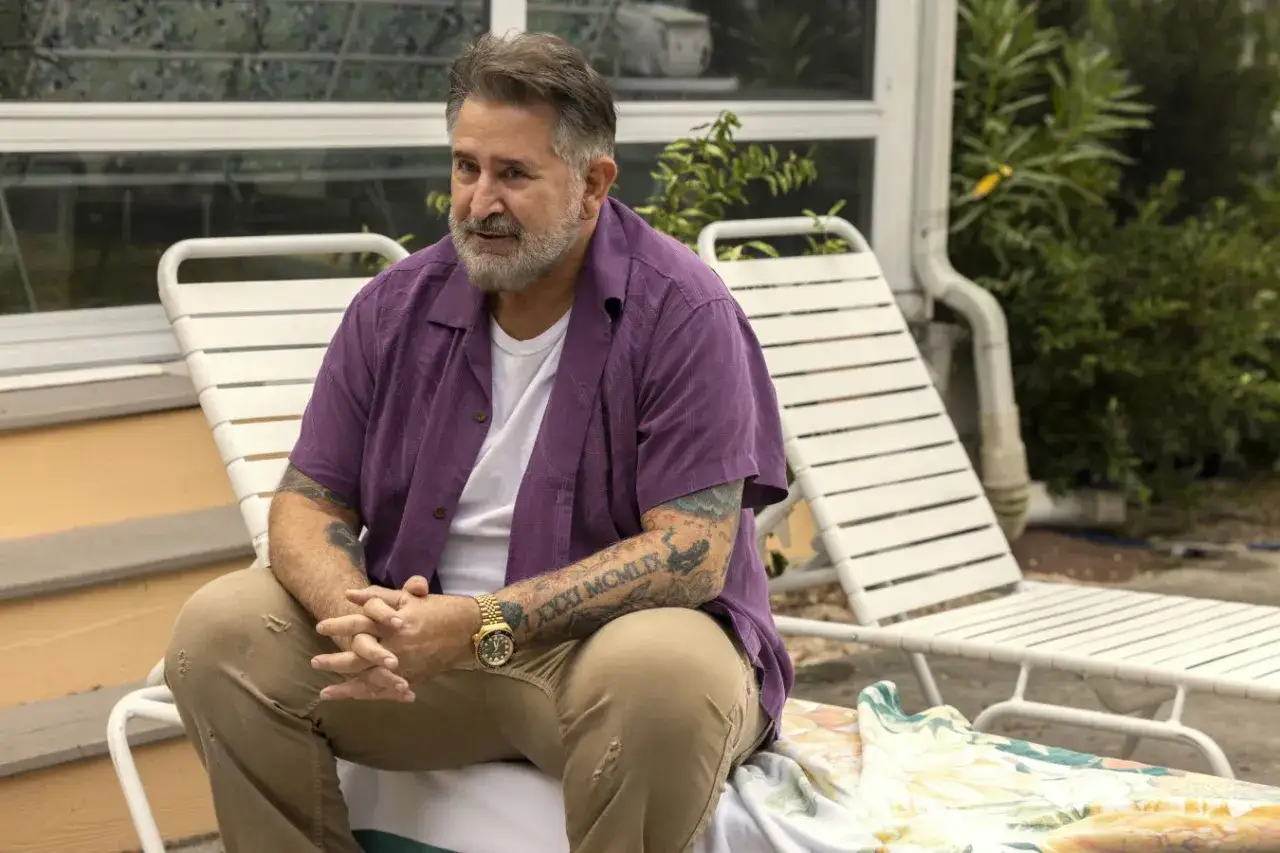 Facet z Florydy, z tatuażami na ramionach, siedzi na leżaku w purpurowej koszuli i beżowych spodniach.