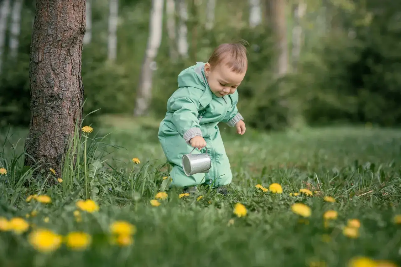 Małe dziecko w zielonym kombinezonie bawi się konewką wśród traw i żółtych kwiatów.