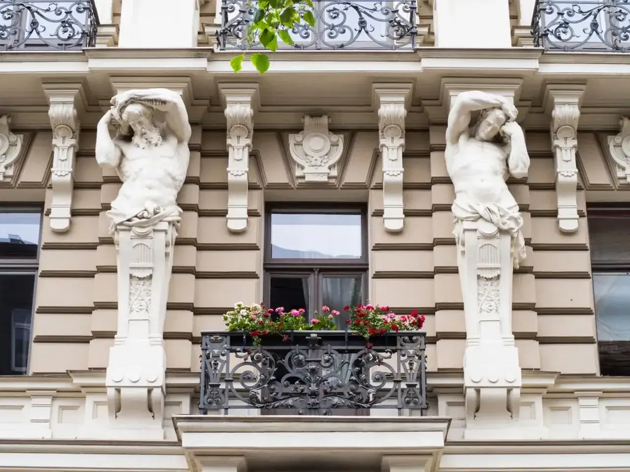 Dwie rzeźby atlantesów podtrzymują balkon w stylu secesyjnym. Kwiaty dodają koloru tej architekturze.