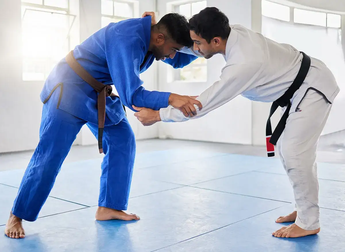 Dwóch mężczyzn w kimonach do BJJ, jeden w niebieskim z brązowym pasem, drugi w białym z czarnym pasem, ćwiczy chwyty na macie.