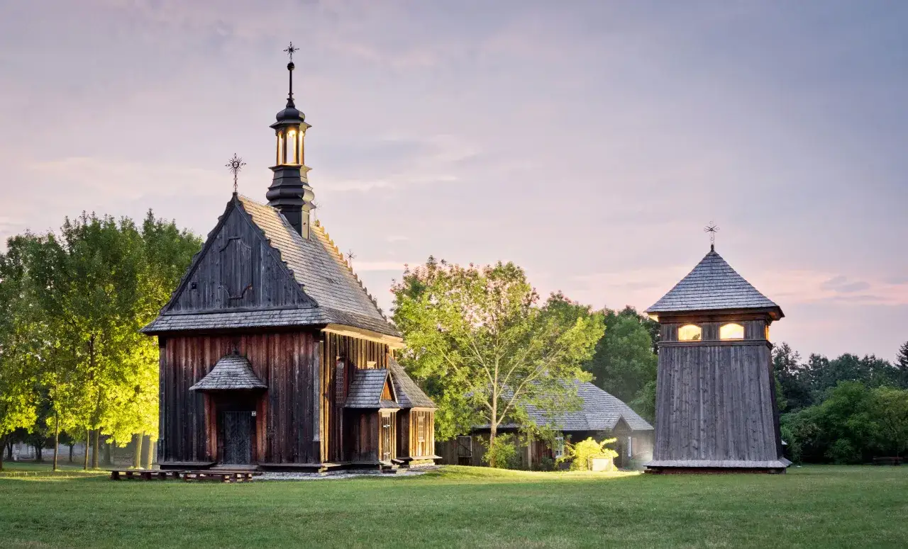 Drewniana cerkiew i dzwonnica na tle zieleni i zachodzącego słońca.