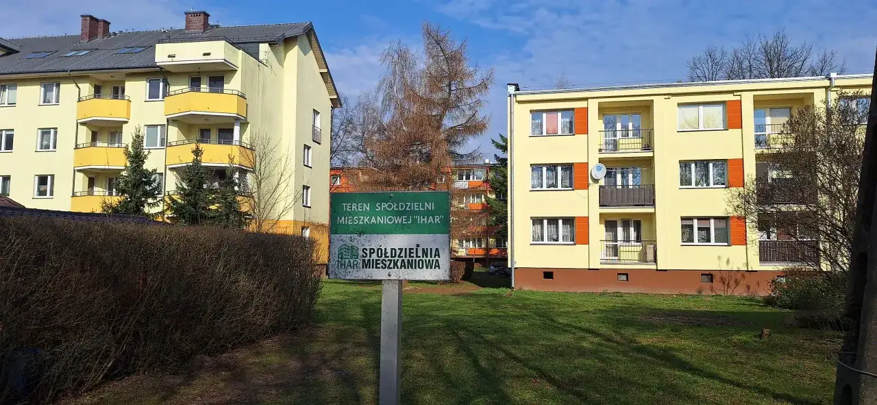 Zielony znak z napisem "TEREN SPÓŁDZIELNI MIESZKANIOWEJ 'IHAR'" przed żółtymi blokami mieszkalnymi.