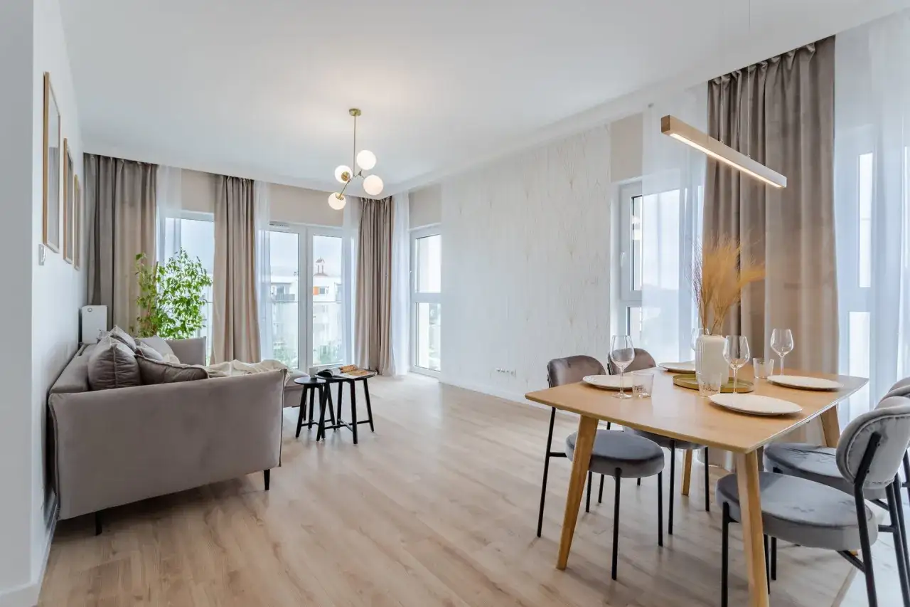Nowoczesny salon z jadalnią. Szara kanapa, drewniany stół z krzesłami, ozdobna ściana i okna. Kto to? Flipper, bo tak nazywa się ten styl.