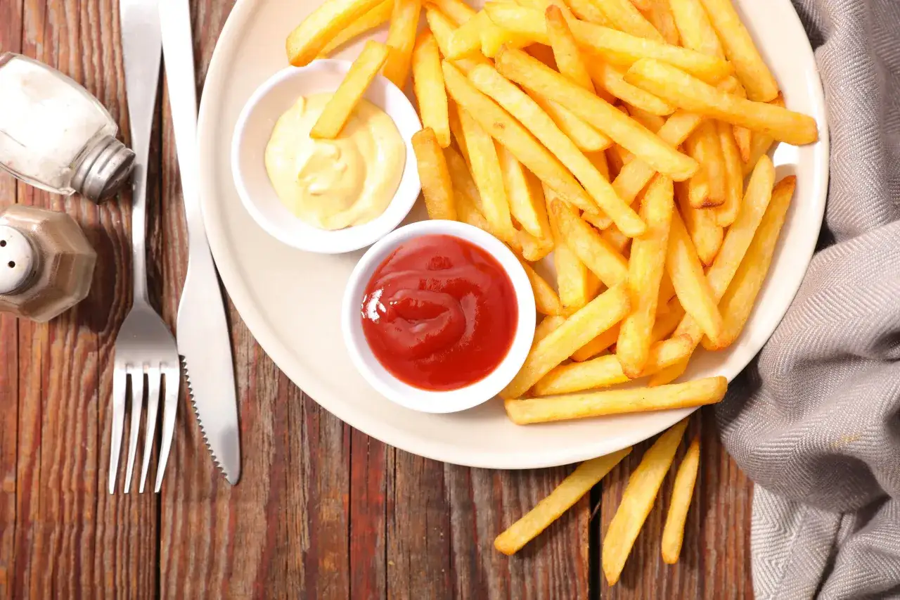 Talerz ze złotymi frytkami, ketchupem, majonezem, solniczką i sztućcami na drewnianym tle.