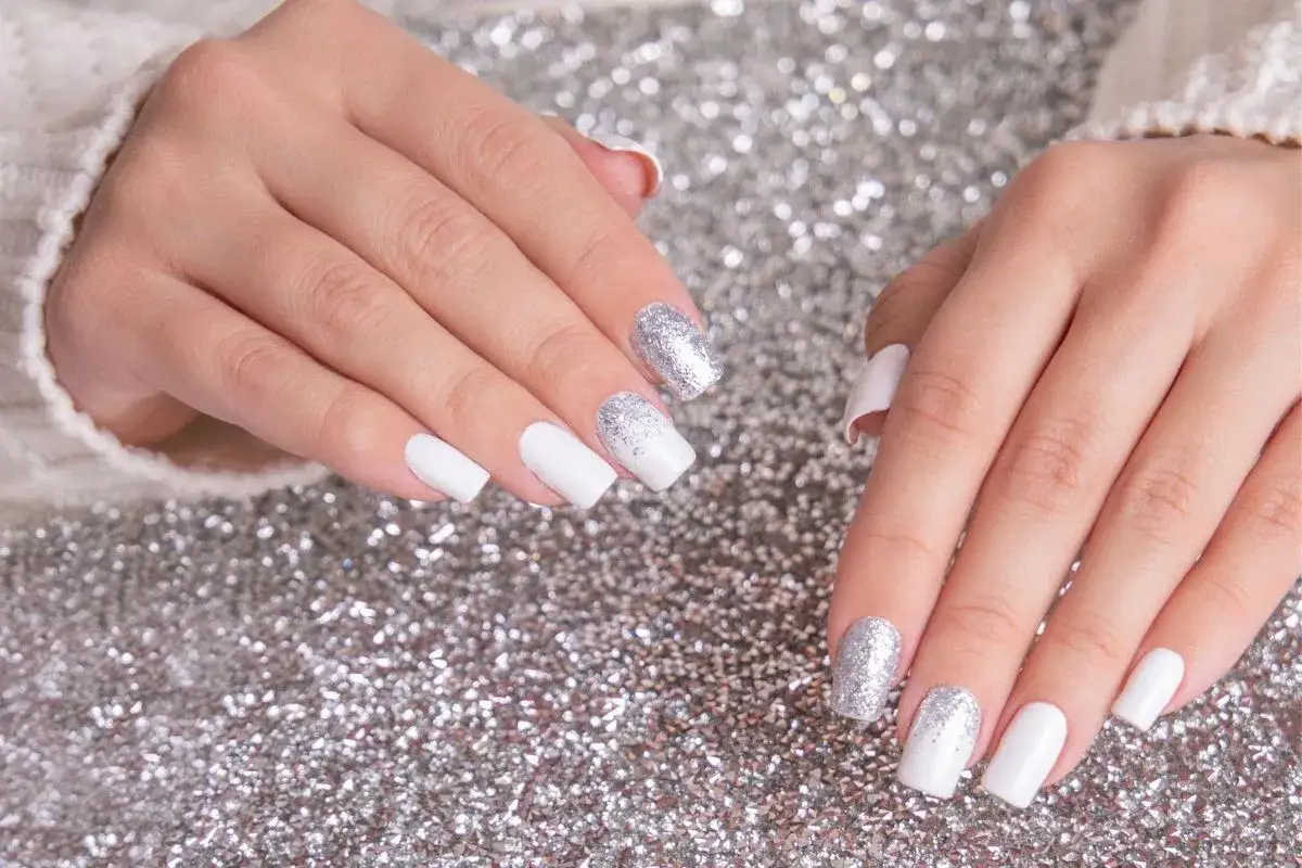 Dłonie z białym french manicure i brokatem na paznokciach, na tle srebrnego brokatu.