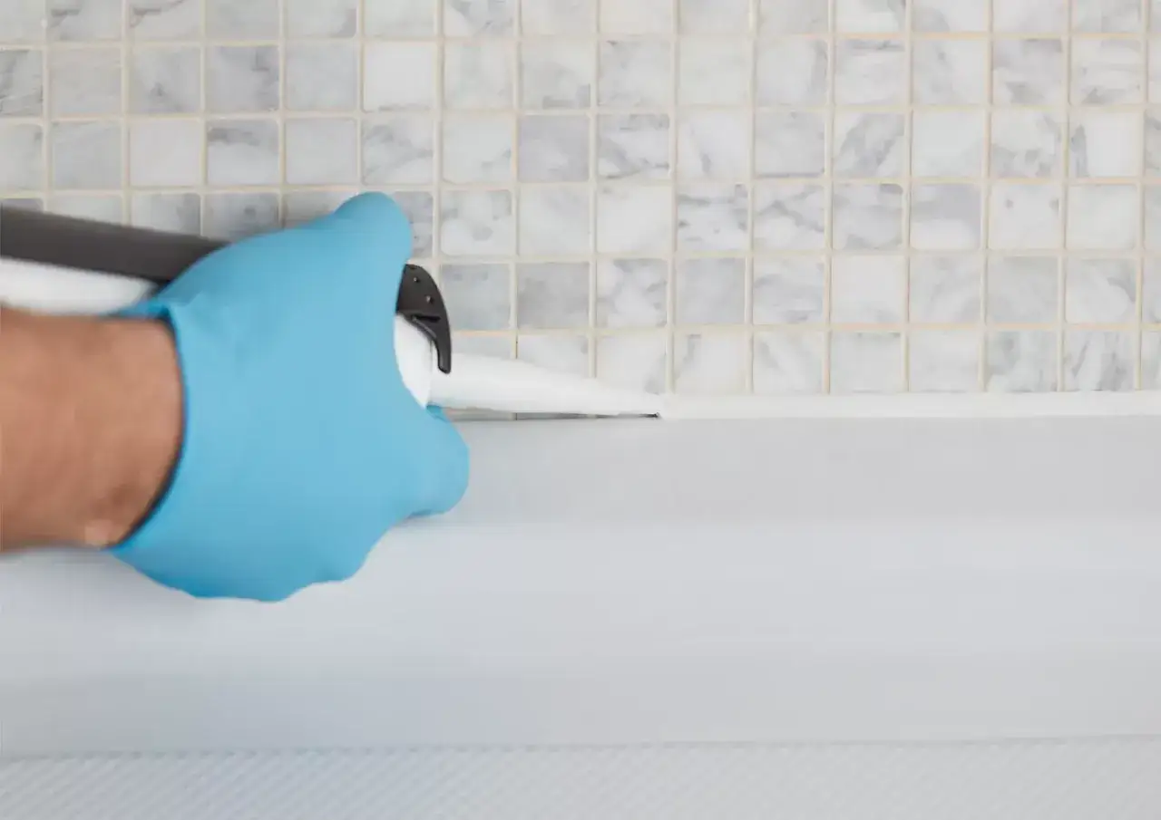 Ręka w niebieskiej rękawiczce aplikuje biały akryl, uszczelniając połączenie wanny z płytkami. Akryl to wszechstronny materiał.