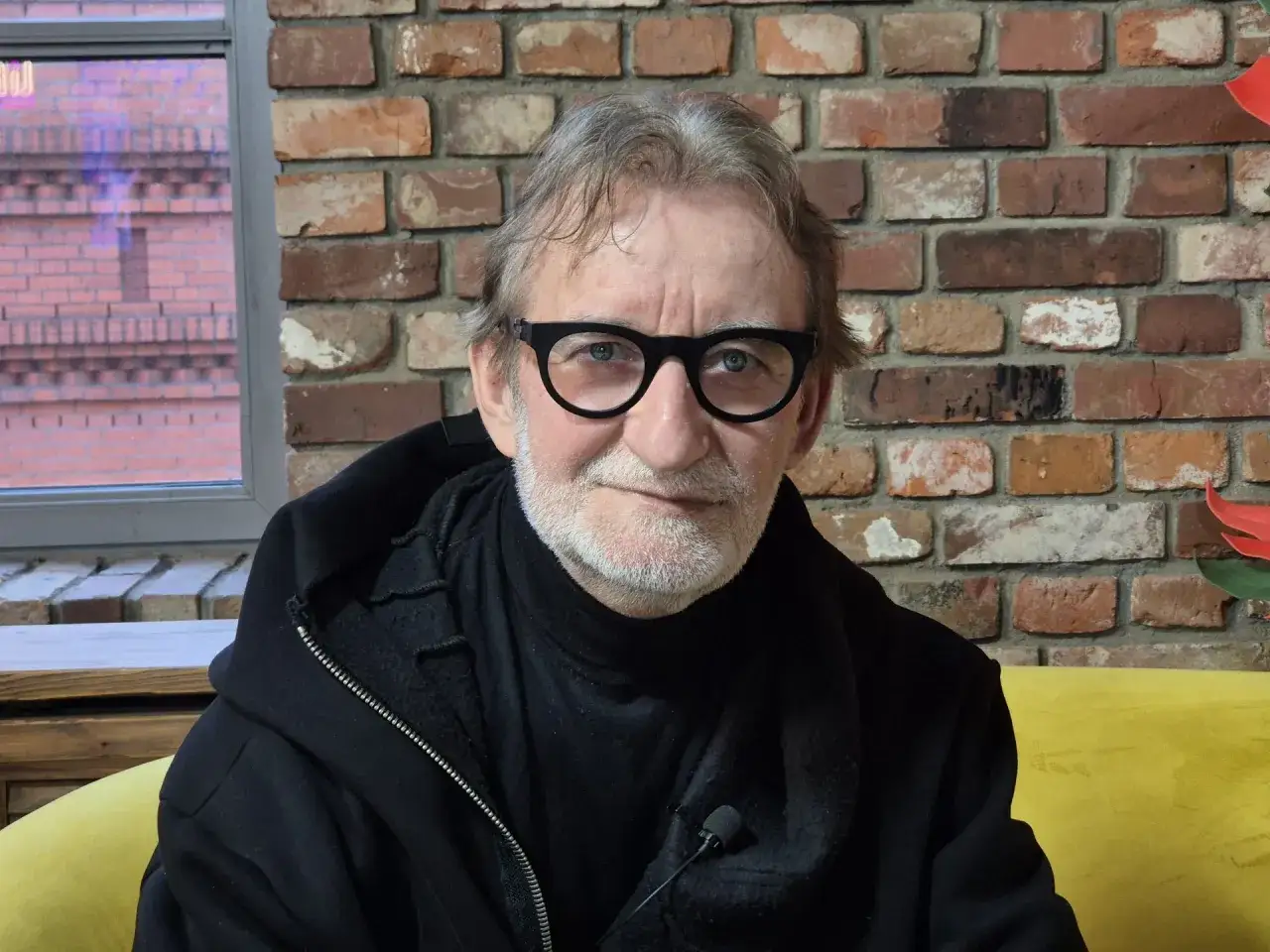 Maciej Mańkowski w okularach, z siwą brodą, w czarnej bluzie na tle ceglanej ściany.