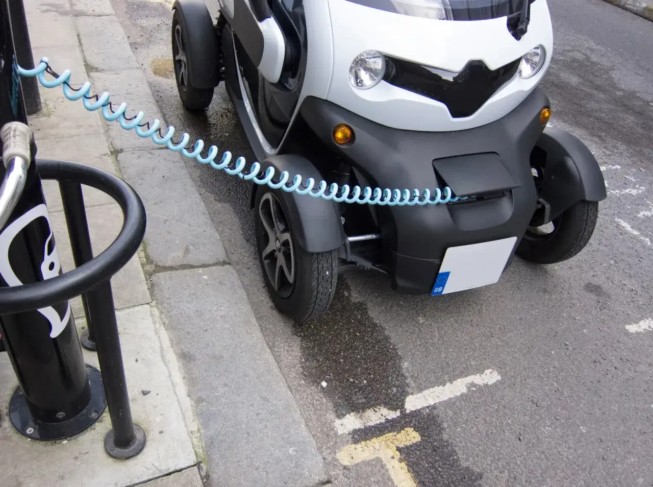 Biały, trójkołowy pojazd elektryczny podłączony do ładowarki. To pokazuje, jak zbudować samochód elektryczny, zaczynając od ładowania.