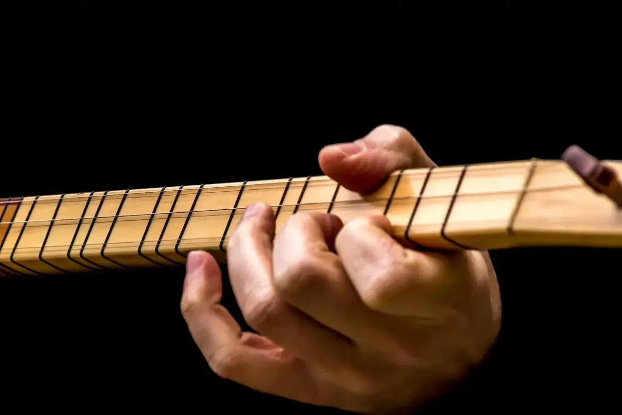 Dłoń gra na instrumencie, dociskając struny. Widoczne są progi i nazwy strun w gitarze.