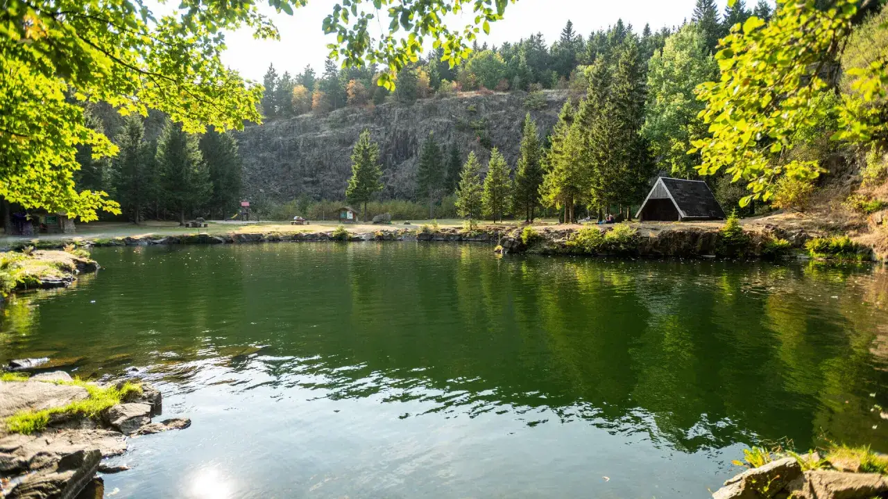 Grüner See mit Bäumen, Felsen und einem kleinen Unterstand am Ufer.
