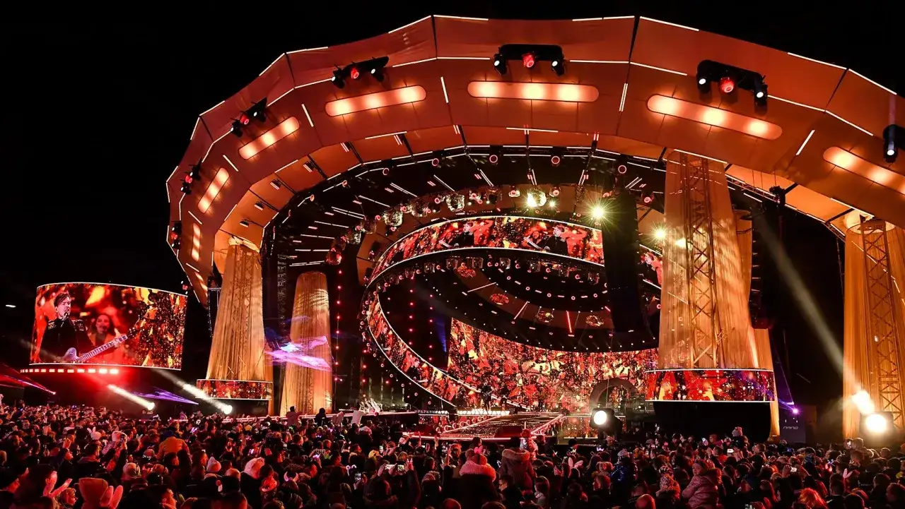 Ogromna scena z ekranami LED i tłumem ludzi. To prawdopodobnie koncert sylwestrowy w Warszawie, pełen świateł i emocji.