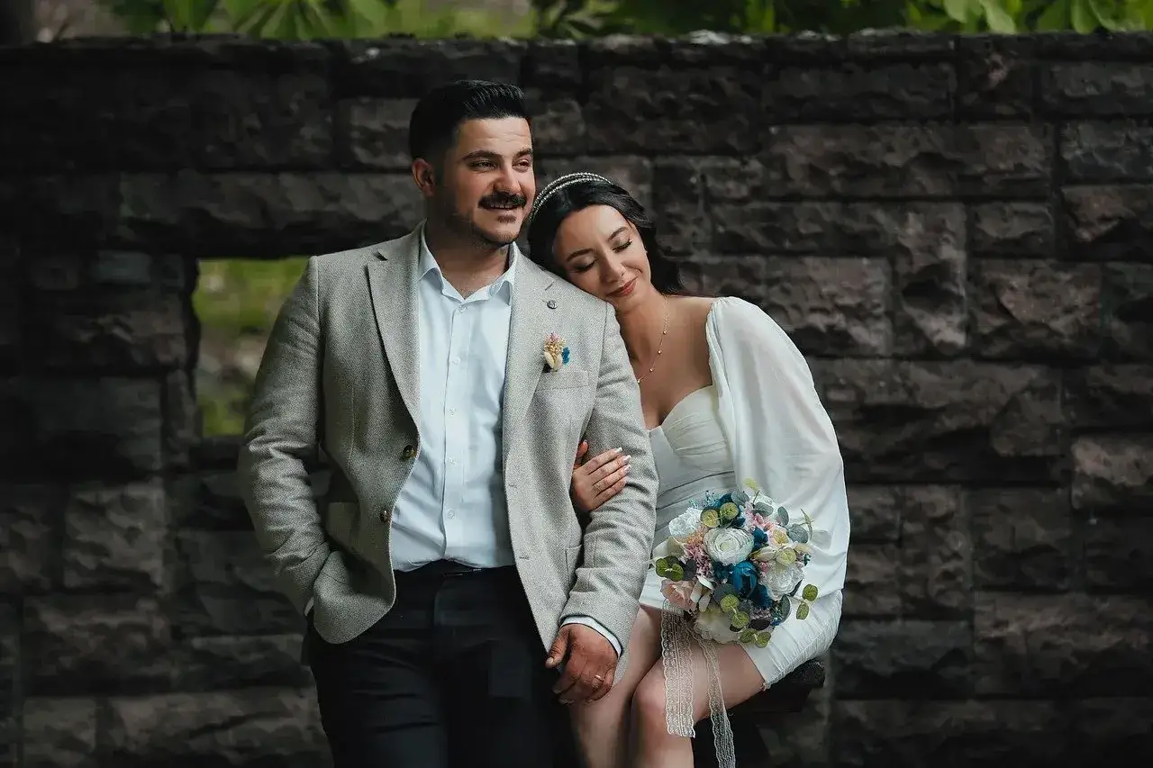 Para młoda w dniu ślubu, ona opiera głowę na jego ramieniu, trzyma bukiet.