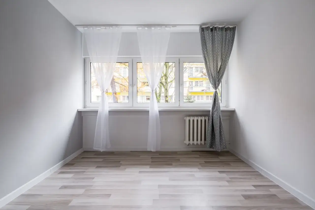 Biały karnisz sufitowy zamontowany nad oknem, na którym wiszą zwiewne firanki i zasłona.