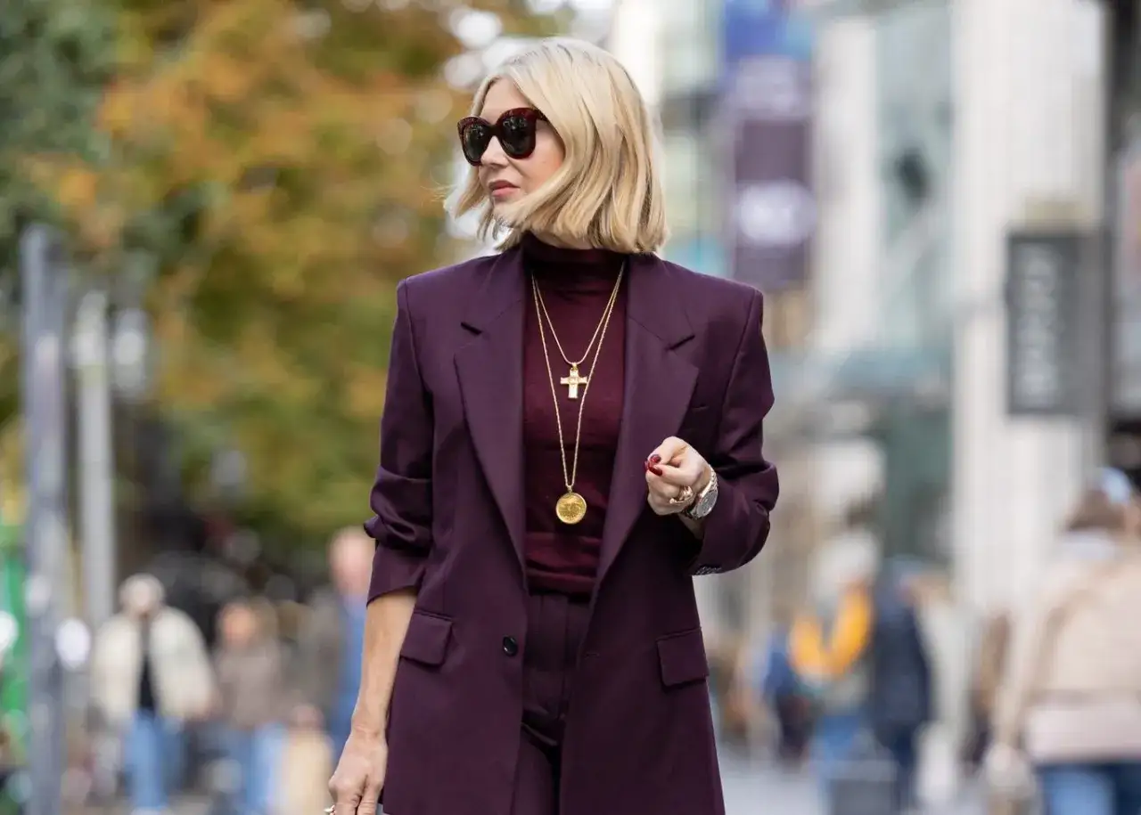 Elegancka kobieta w garniturze w kolorze burgundowym, z złotym krzyżykiem na szyi. Ten kolor pasuje do każdej pewnej siebie kobiety.