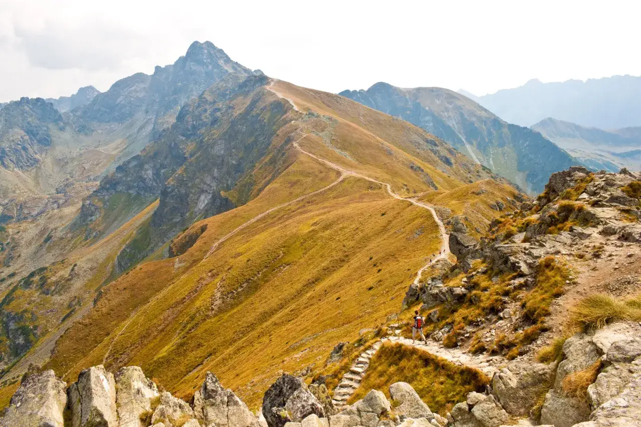 Szlak turystyczny wije się pośród skalistych zboczy, prowadząc ku majestatycznym, jesiennym szczytom. W oddali widać kolejne pasma górskie.
