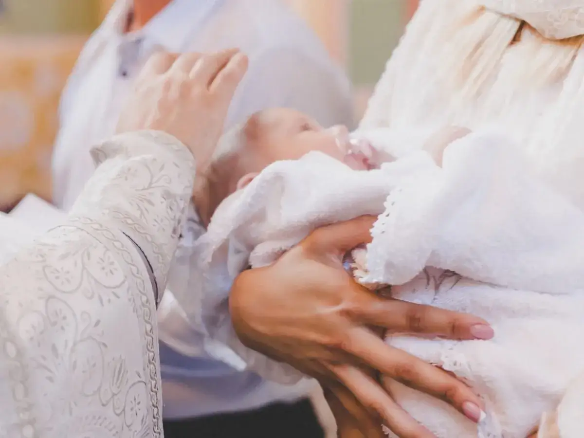 Dziecko owinięte w biały ręcznik, trzymane na rękach. Może to prezent od cioci na chrzest.