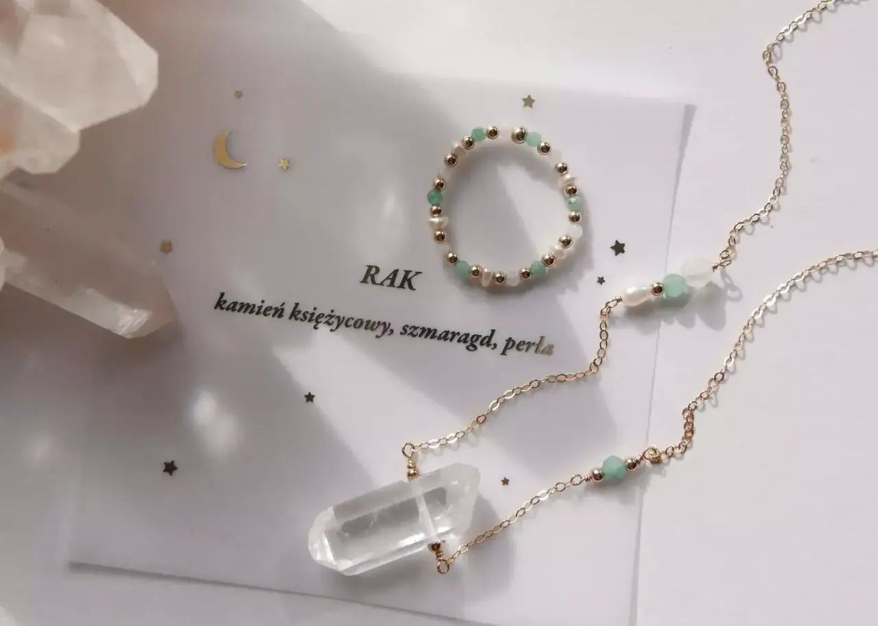 Złota biżuteria z kryształem, perłami i zielonymi kamieniami. Napis "Rak" i "kamień księżycowy, szmaragd, perła" sugerują, że to kamień zodiakalny dla Raka.