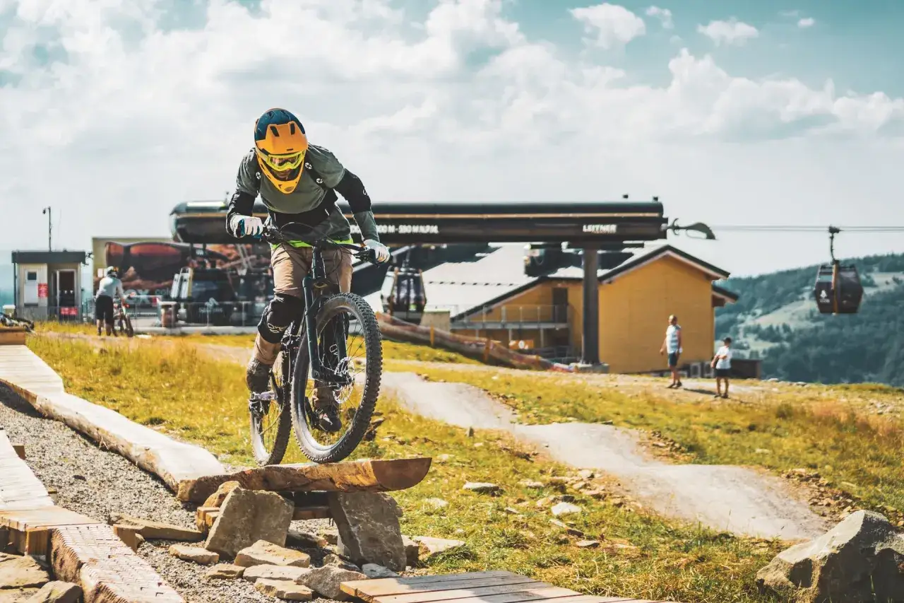Rowerzysta pokonuje przeszkodę na drewnianej kładce w bike park Bielsko Biała. W tle kolejka linowa i budynek stacji.