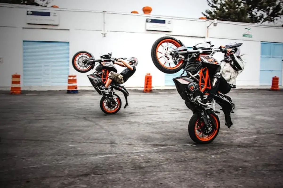 Dwóch motocyklistów wykonuje wheelie na swoich pomarańczowych motocyklach.