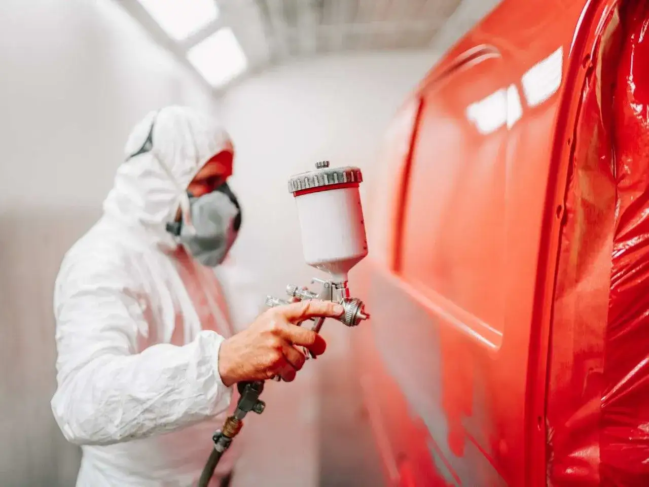 Lakiernik w kombinezonie ochronnym używa pistoletu z **zbiornikiem do pistoletu lakierniczego**, by nadać karoserii lśniący, czerwony kolor.