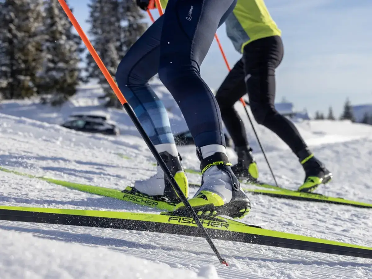 Dwie osoby na nartach biegowych pokonują ośnieżony stok. Słońce odbija się od ich nart Fischer.