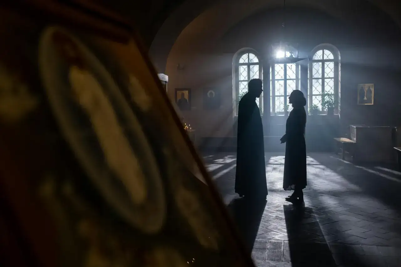 Sylwetki księdza i kobiety rozmawiających w kościele, w świetle wpadającym przez okna.