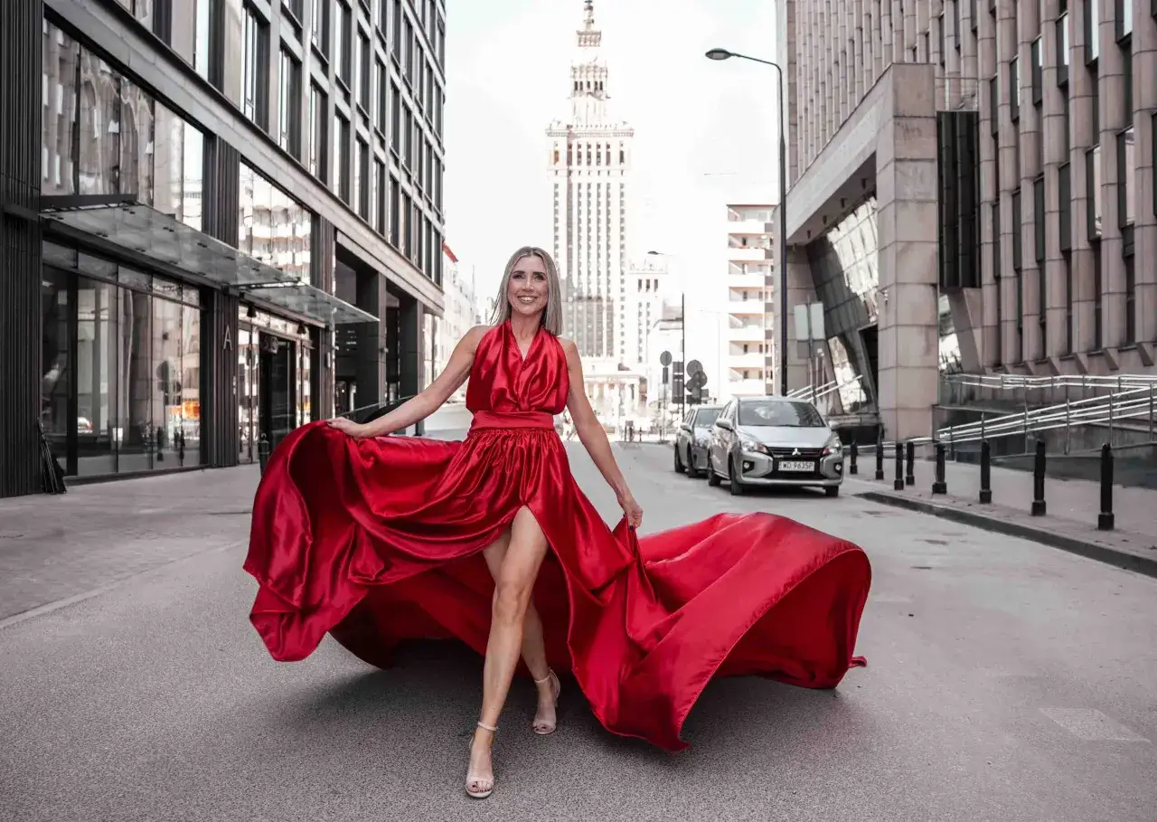 Kobieta w czerwonej sukni z rozcięciem na nodze idzie ulicą, trzymając materiał sukni.