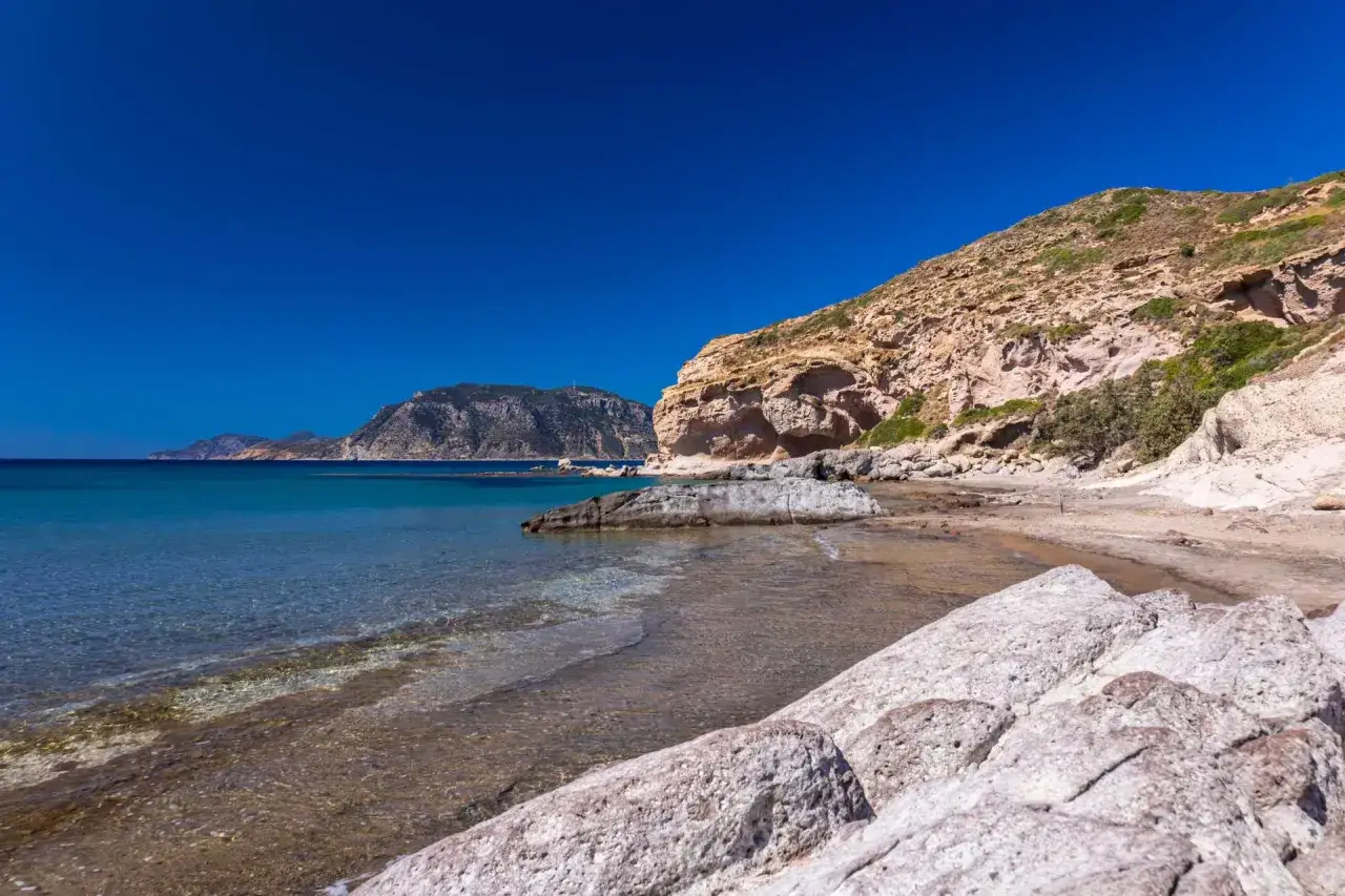 Majowa pogoda na Kos: skalista plaża z turkusową wodą i łagodnymi falami, w tle góry.