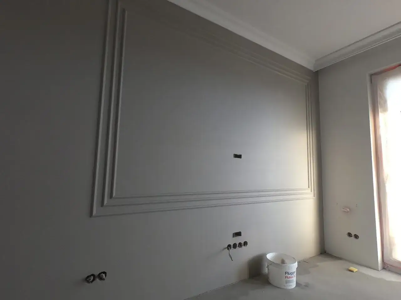 Ściana z ozdobnymi listwami, przygotowana do malowania. Widać wiadro z farbą, co sugeruje, że zaraz nastąpi malowanie agregatem bez smug.