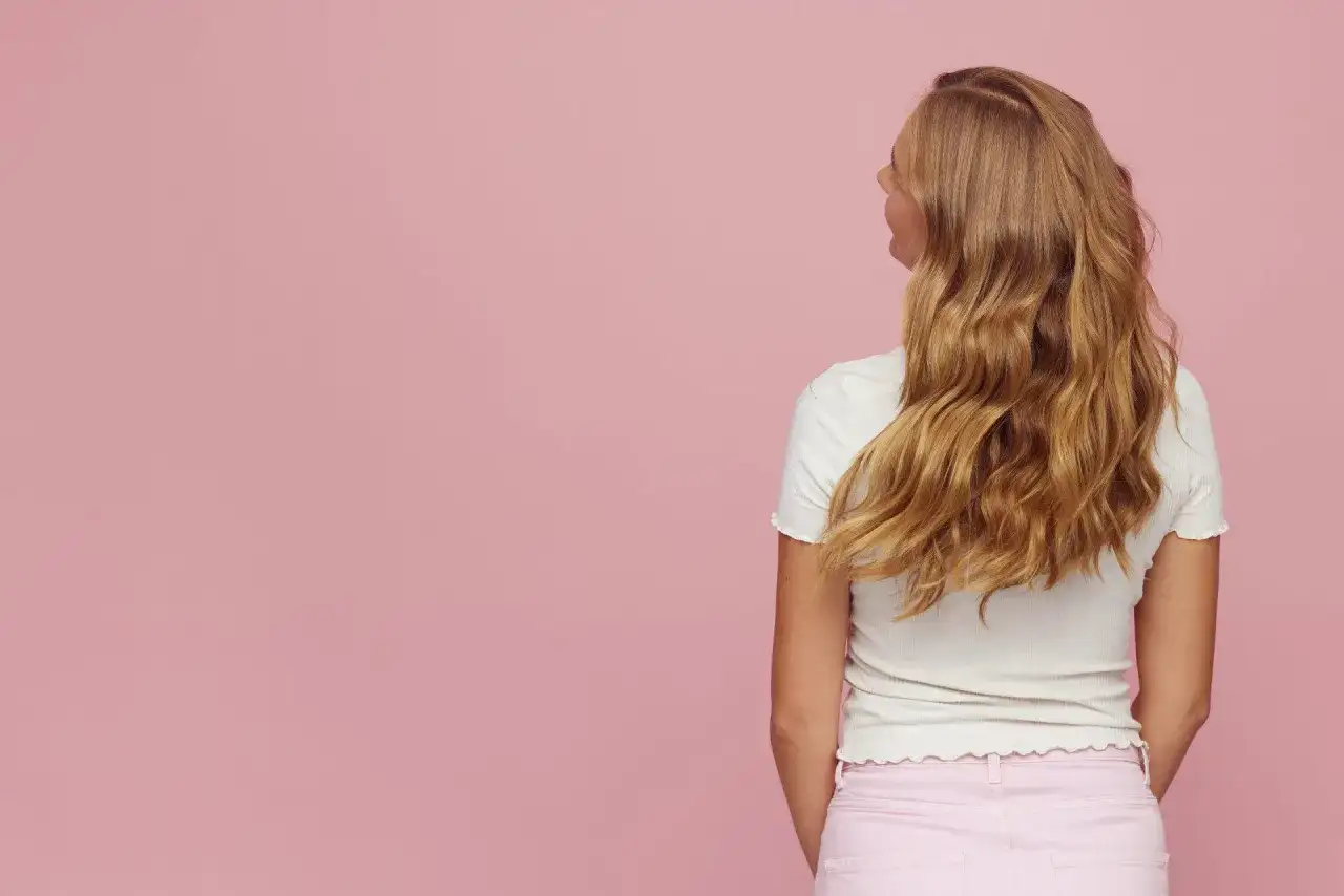 Blond włosy z efektem butterfly cut, falujące i lśniące, na tle pudrowego różu.