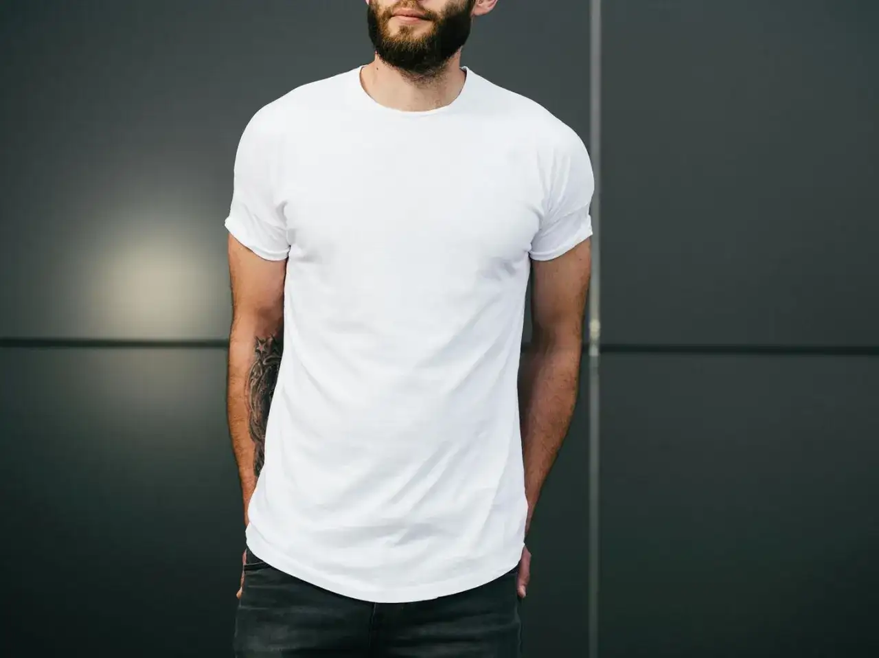 Mann mit Bart trägt weißes T-Shirt und Jeans, Hände in den Taschen.