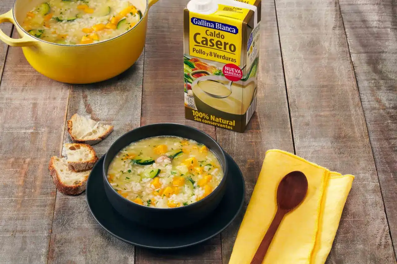 Un tazón humeante de sopa de arroz con calabacín y zanahoria, junto a pan rústico y un cartón de caldo Gallina Blanca.