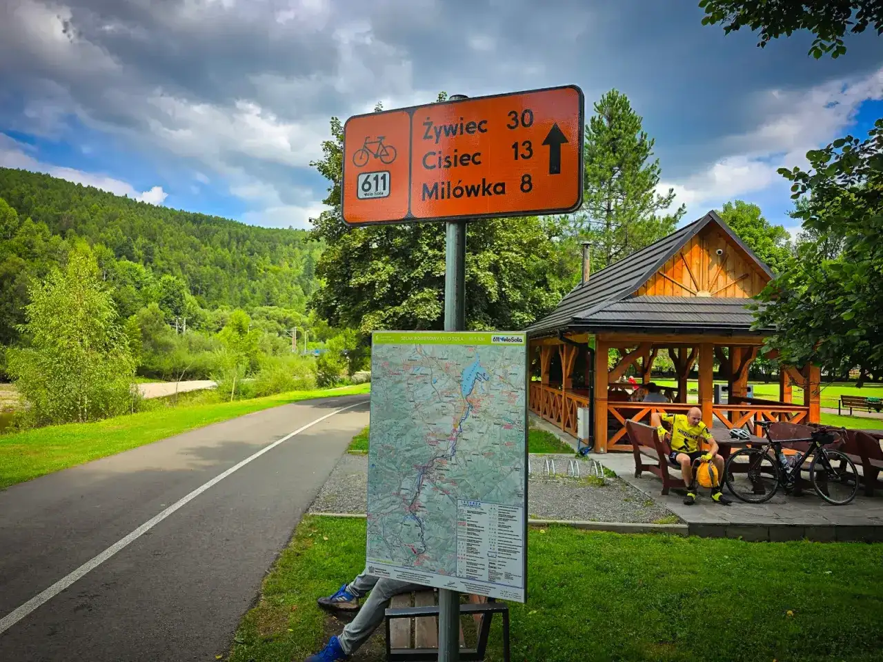 Znak szlaku rowerowego Velo Sola z odległościami do Żywca, Ciśca i Milówki. Obok mapa i wiata.