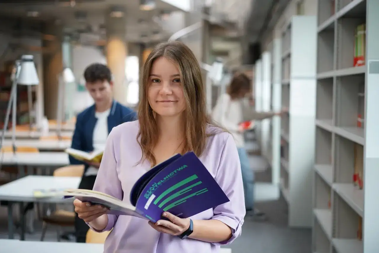 Studentka z uśmiechem trzyma broszurę, być może dotyczącą stypendium socjalnego. W tle inni studenci w bibliotece.