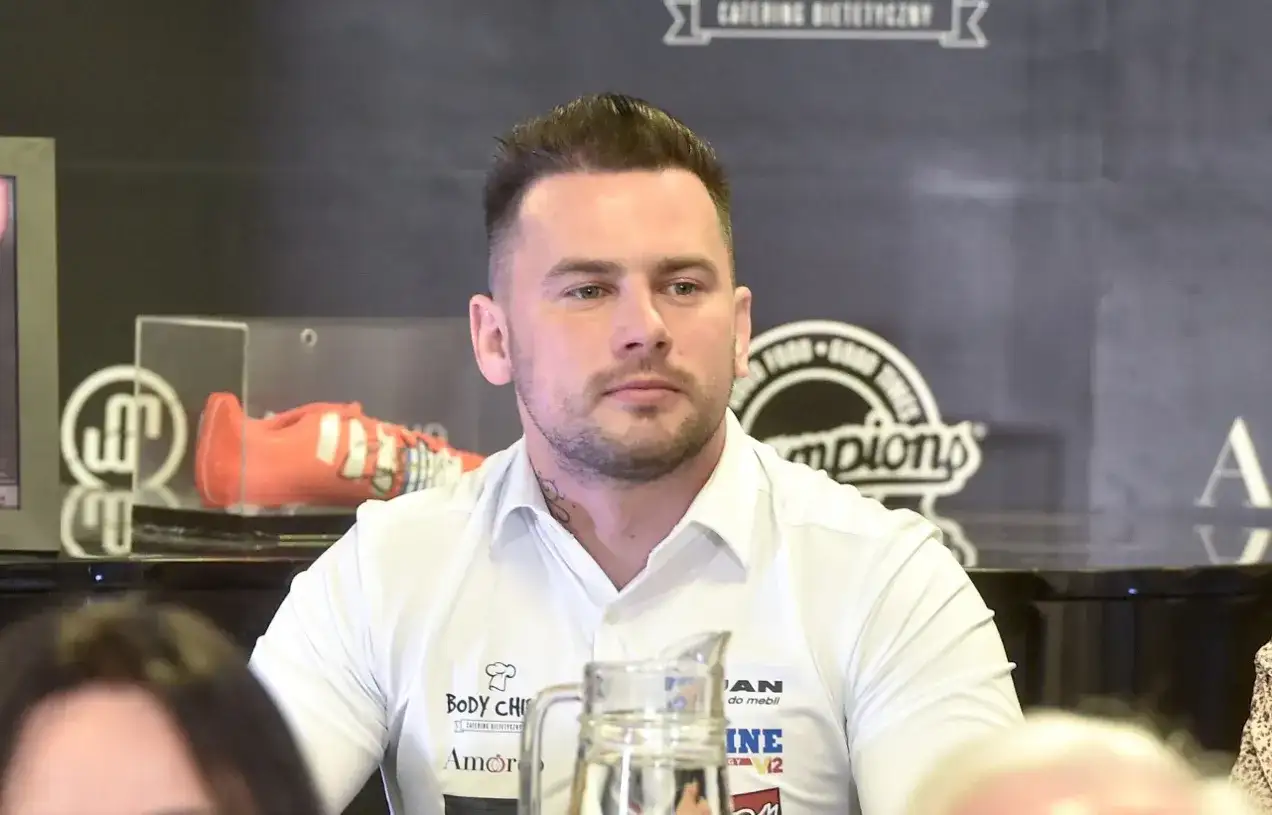 Grzegorz Collins, młody mężczyzna w białej koszuli, z tatuażem na szyi, siedzi przy stole. W tle widoczny pomarańczowy but sportowy.