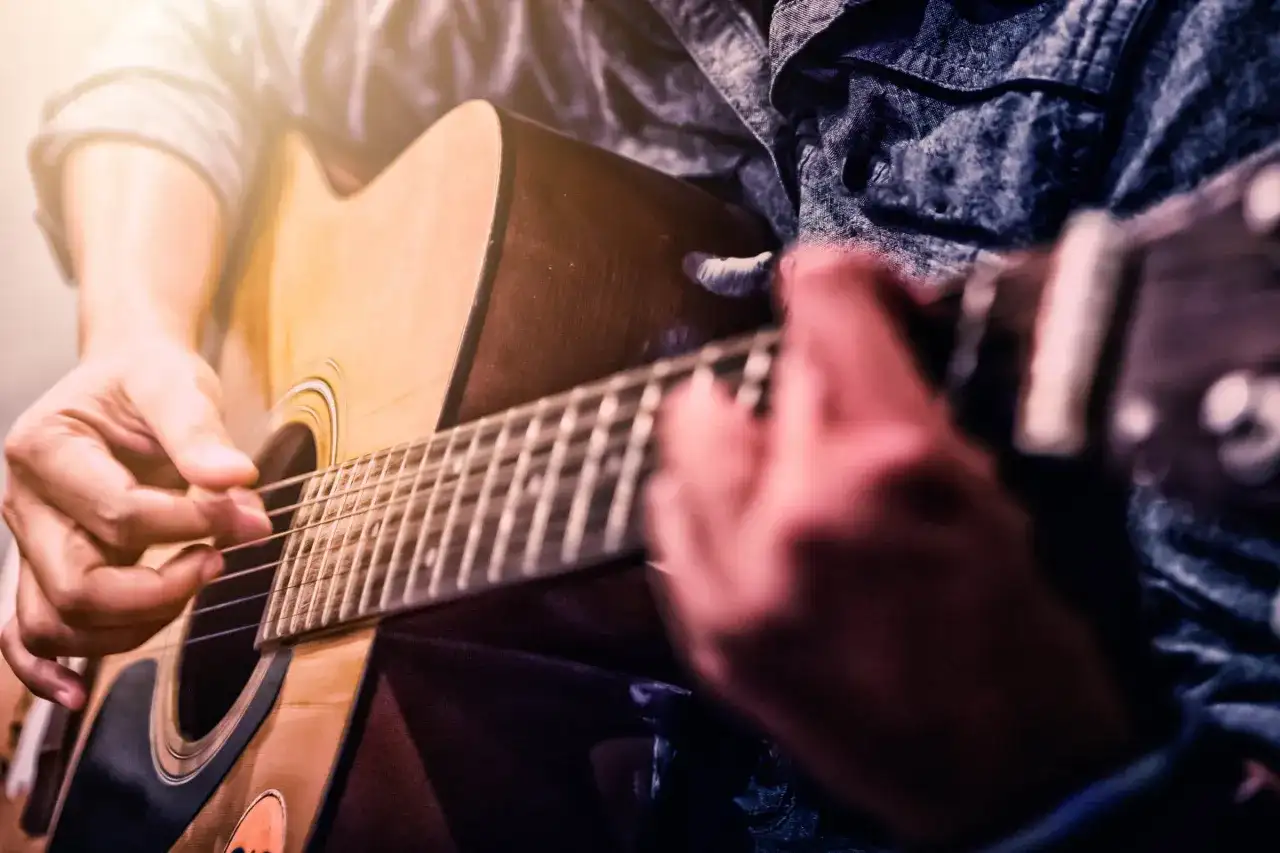 Dłonie grają na gitarze akustycznej. W tle widać fragmenty odzieży, przypominające dżinsową koszulę. To piękne instrumenty muzyczne, które tworzą niepowtarzalny dźwięk.