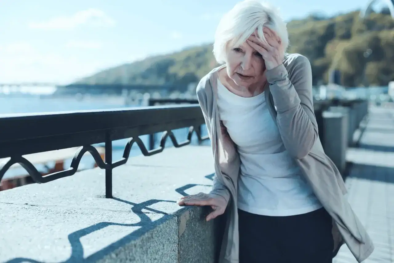 Starsza kobieta z bólem głowy, być może potrzebuje cinnarizinum, opiera się o barierkę nad wodą.