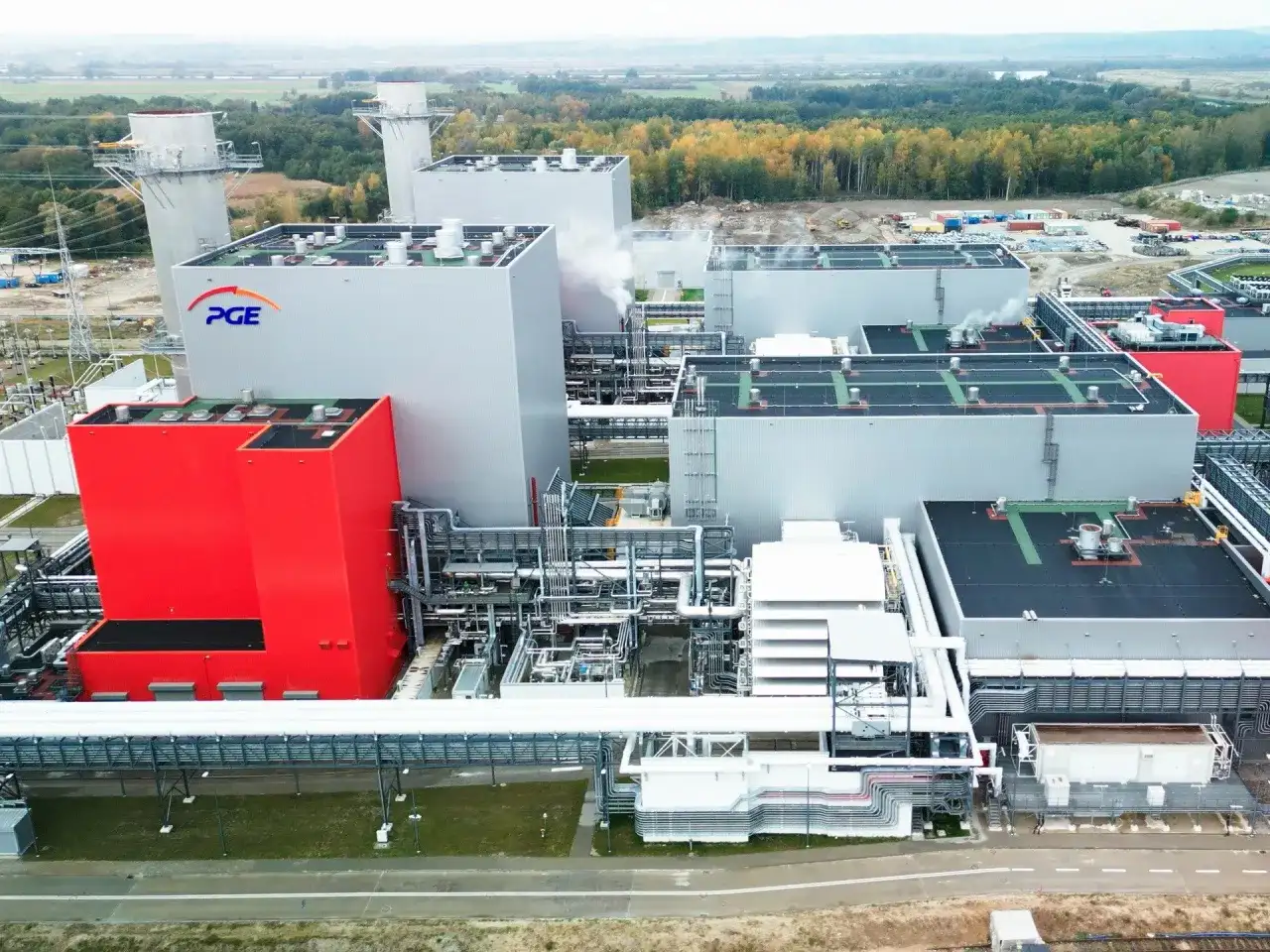 Nowoczesna elektrownia PGE z czerwonym budynkiem i białymi blokami, w tle las i kominy.