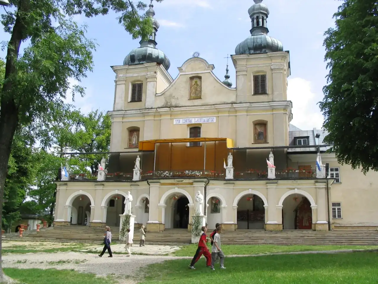 Kalwaria Pacławska: Sanktuarium z kopułami i tarasem, gdzie pielgrzymi spacerują pośród zieleni.