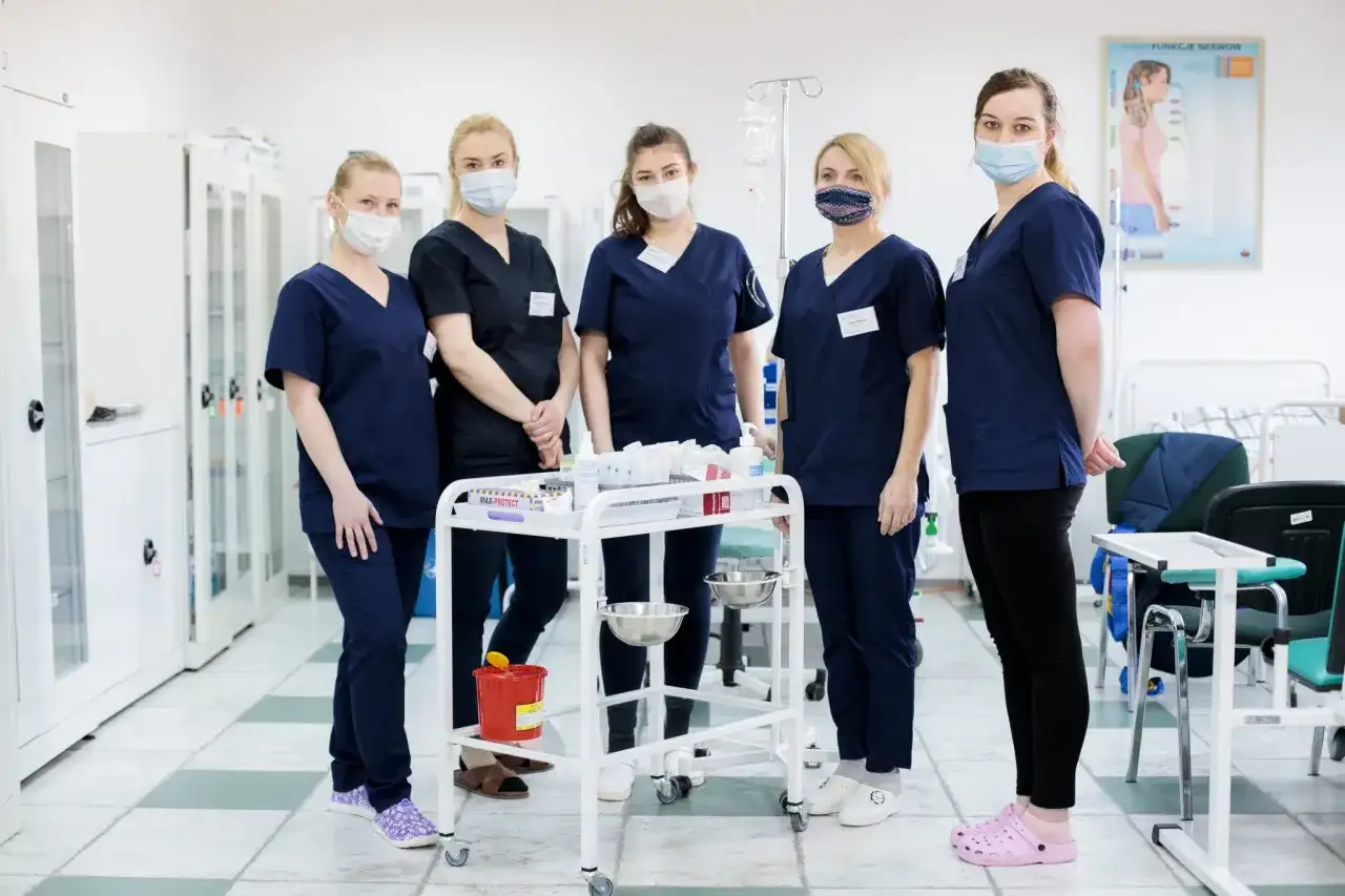 Grupa studentek pielęgniarstwa w maseczkach stoi przy wózku medycznym w sali szpitalnej.