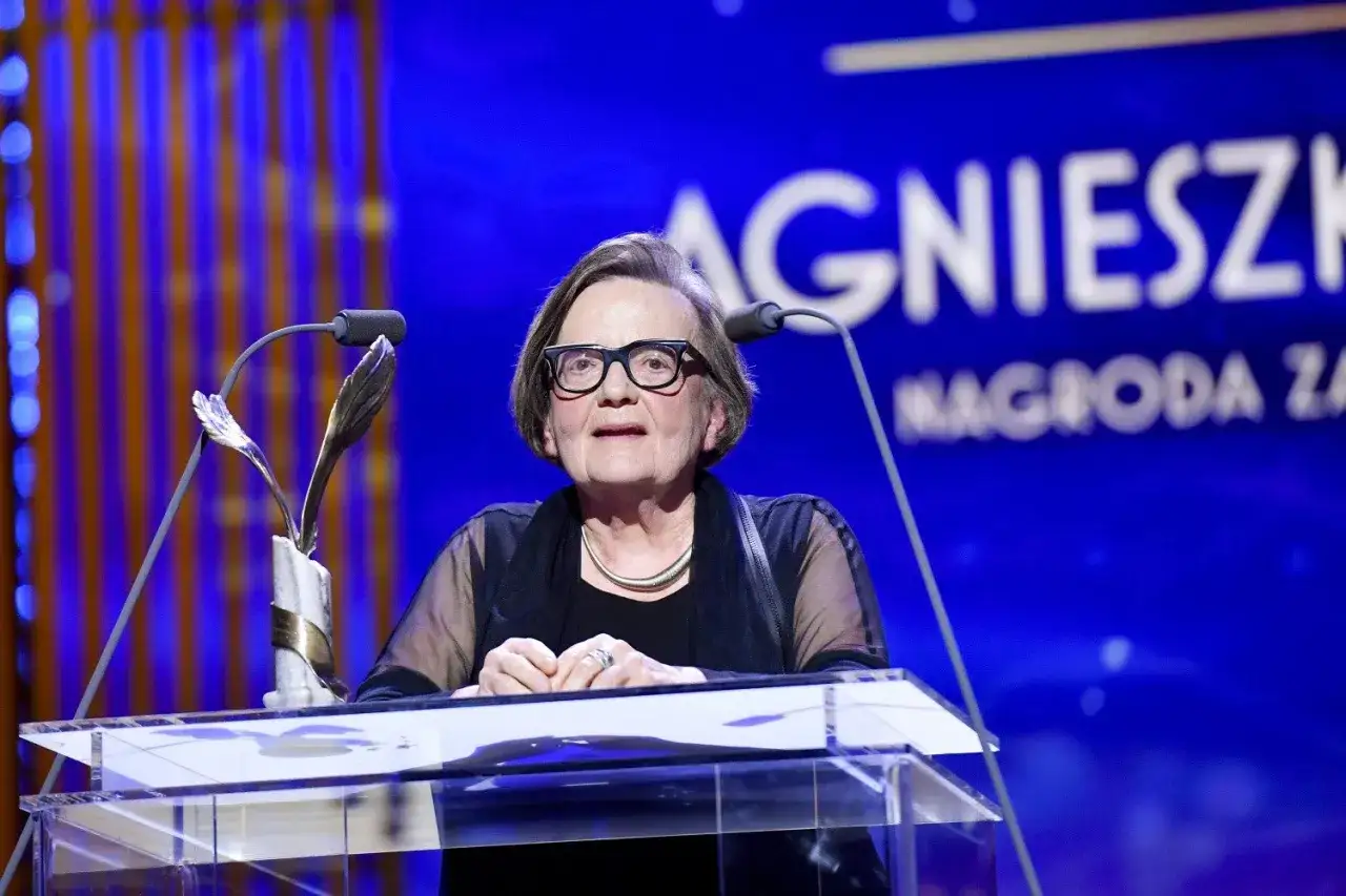 Agnieszka Holland w okularach za mównicą z mikrofonem i statuetką.