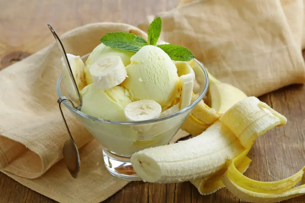 Pyszne lody bananowe w pucharku, ozdobione plasterkami banana i listkiem mięty. Obok leży obrany banan.
