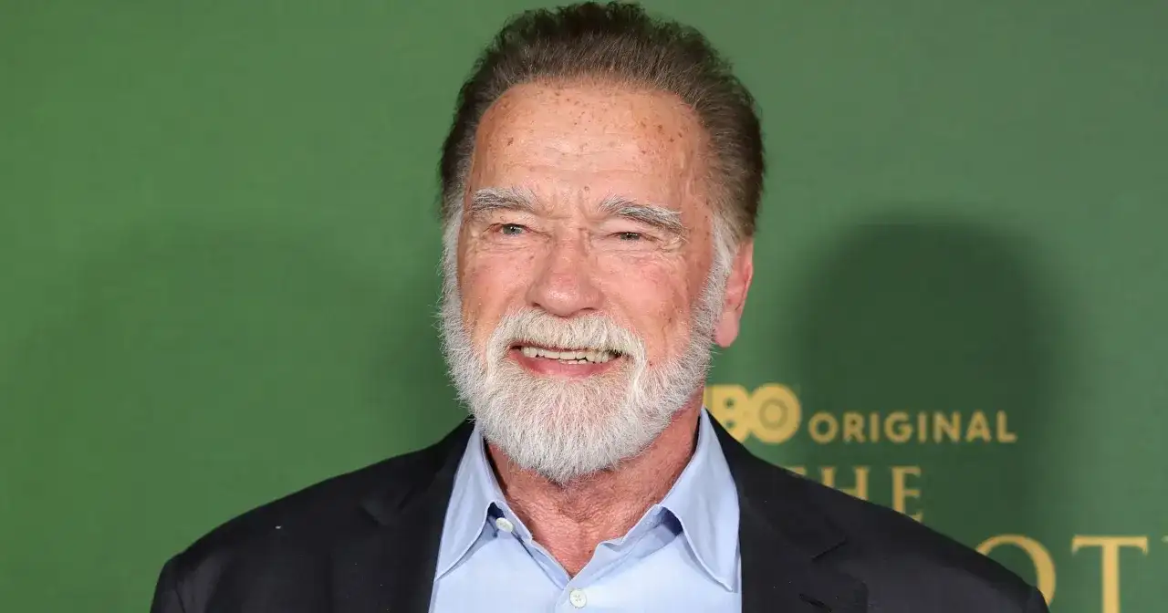 Arnold Schwarzenegger, z siwą brodą i w niebieskiej koszuli, uśmiecha się. W tle zielona ściana z logo HBO.