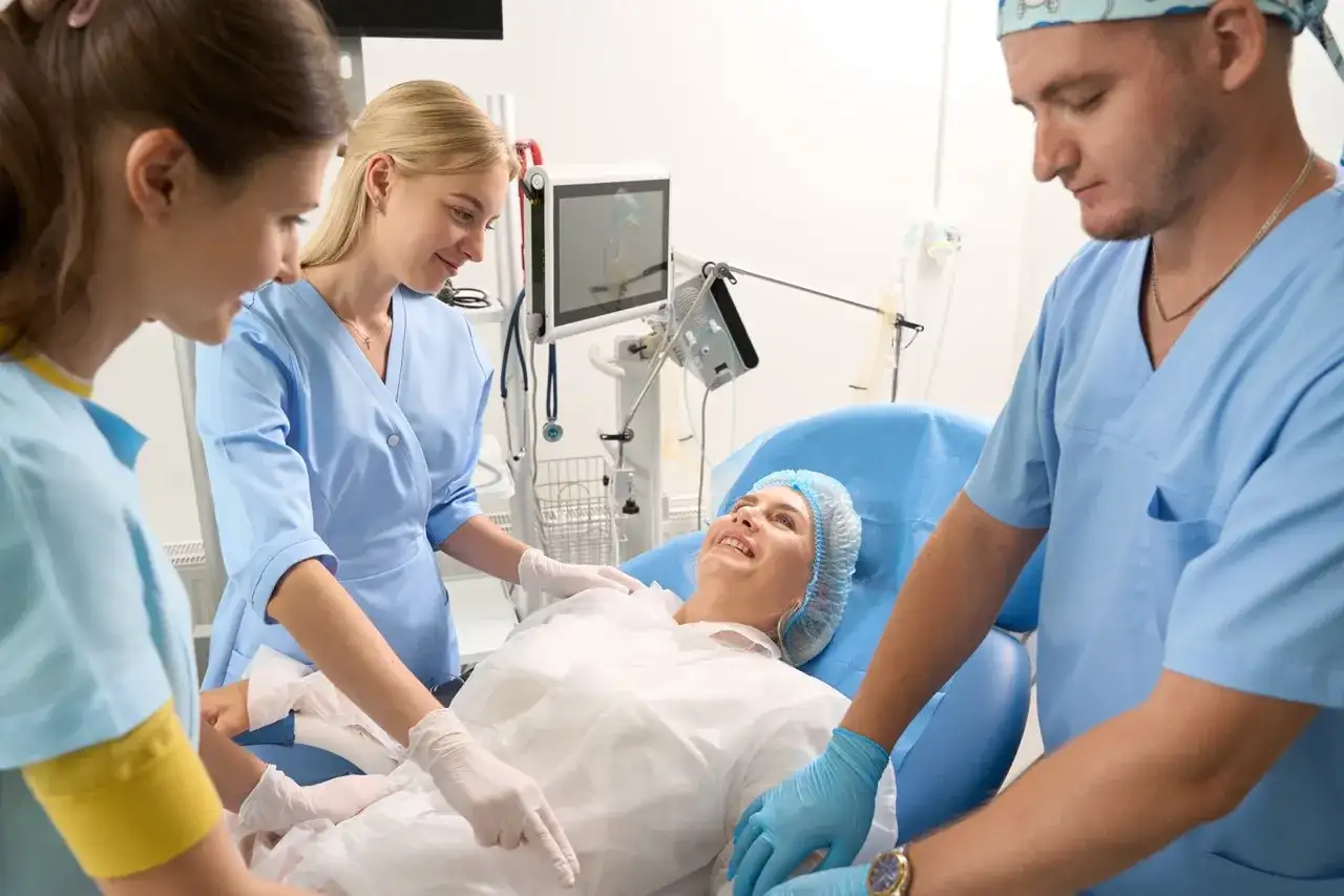 Uśmiechnięta pacjentka w czepku leży na łóżku, otoczona przez zespół medyczny. Opiekun medyczny i dwie pielęgniarki czuwają nad jej komfortem.