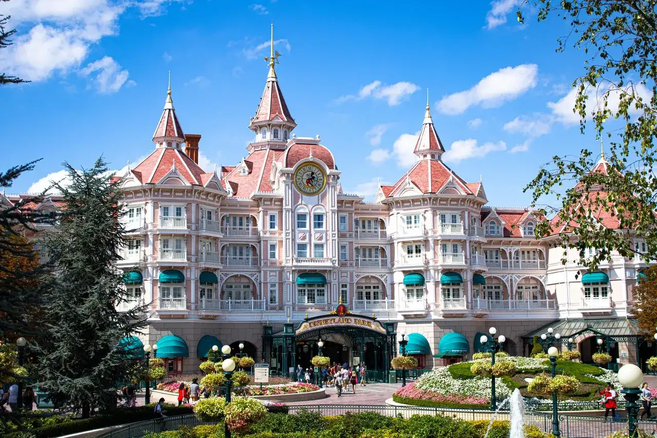 Grand hôtel rose de style victorien avec des tours, un grand cadran d'horloge et des jardins fleuris.