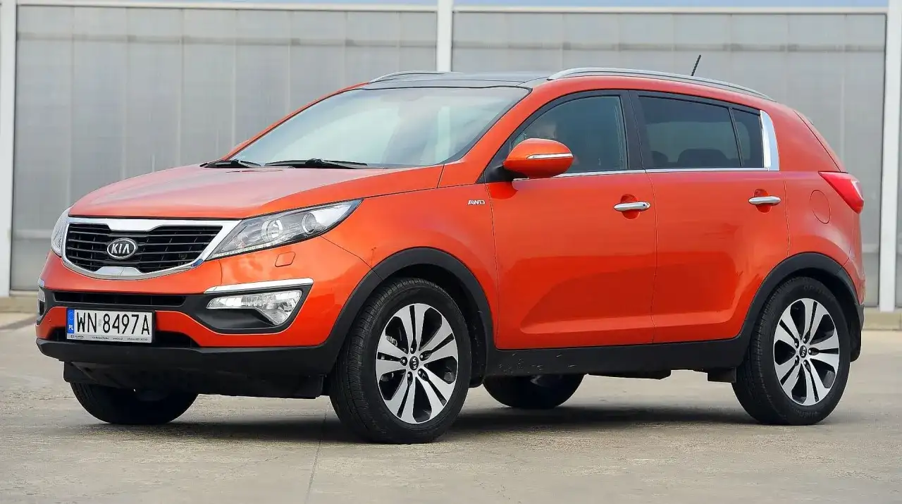 Pomarańczowy Kia Sportage 2015 z napędem AWD, zaparkowany na betonowym placu przed budynkiem z przeszkloną fasadą.