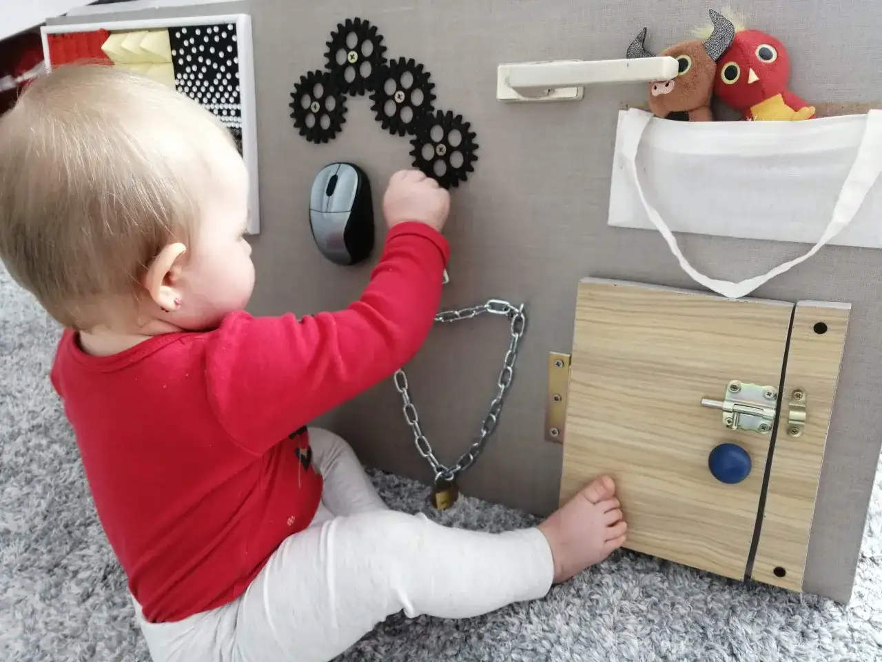 Dziecko bawi się przy tablicy manipulacyjnej diy: zębatki, myszka, zamek, kłódka i pluszaki.