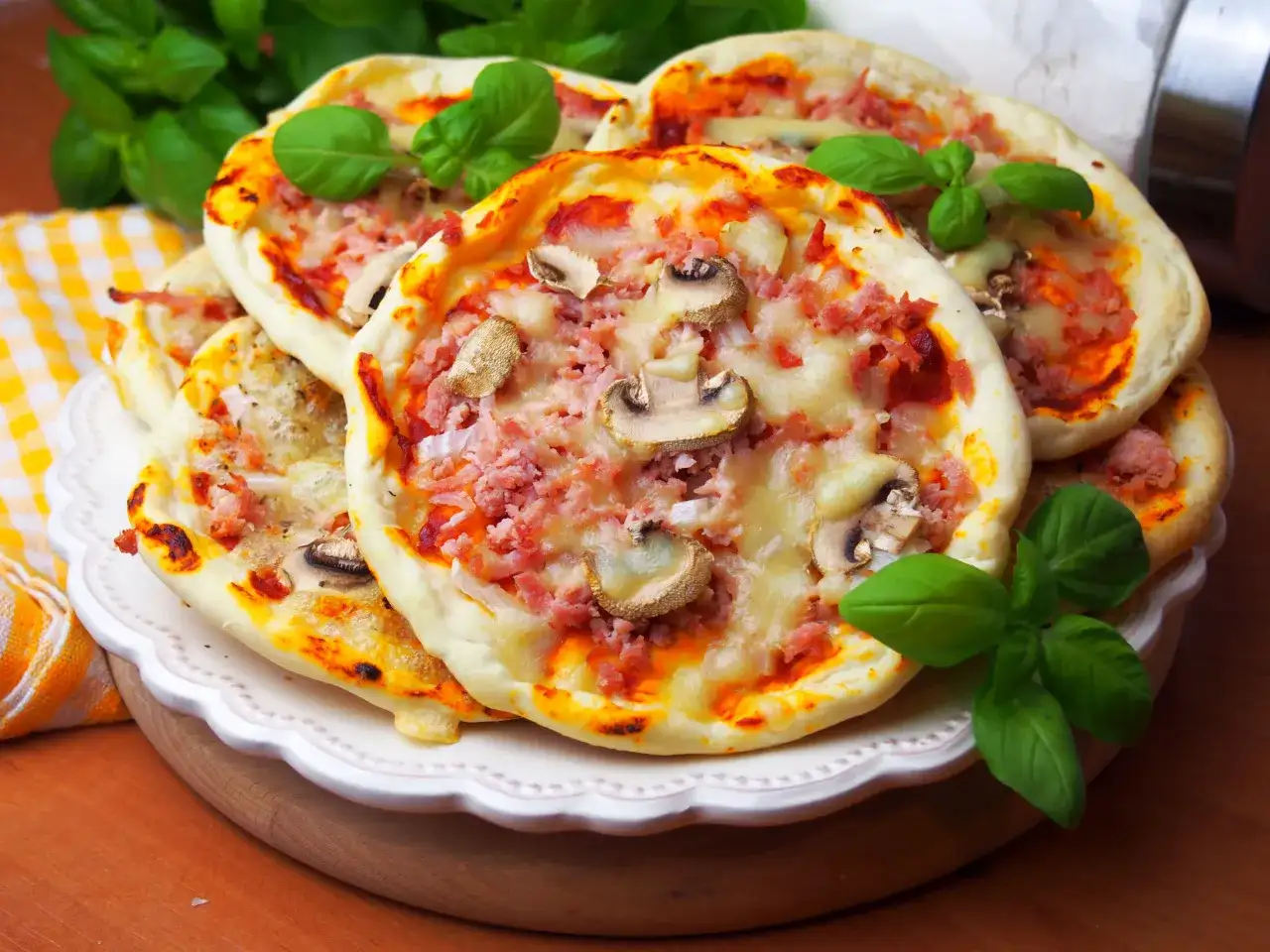 Szybkie mini pizze z szynką i pieczarkami, ozdobione świeżą bazylią, kuszą apetycznym wyglądem.