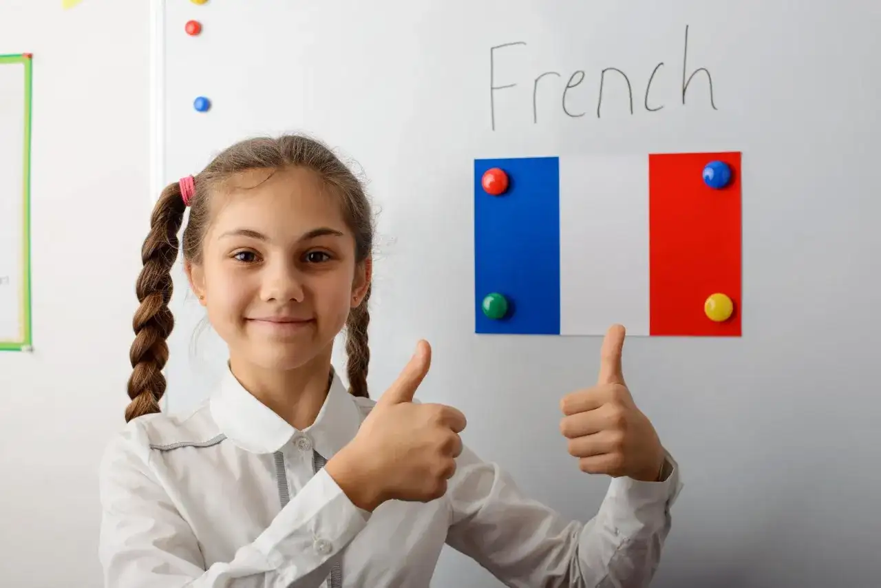 Uśmiechnięta dziewczynka z warkoczami pokazuje kciuki w górę. Na tablicy napis "French" i flaga Francji. Edukacja we Francji ciekawostki.
