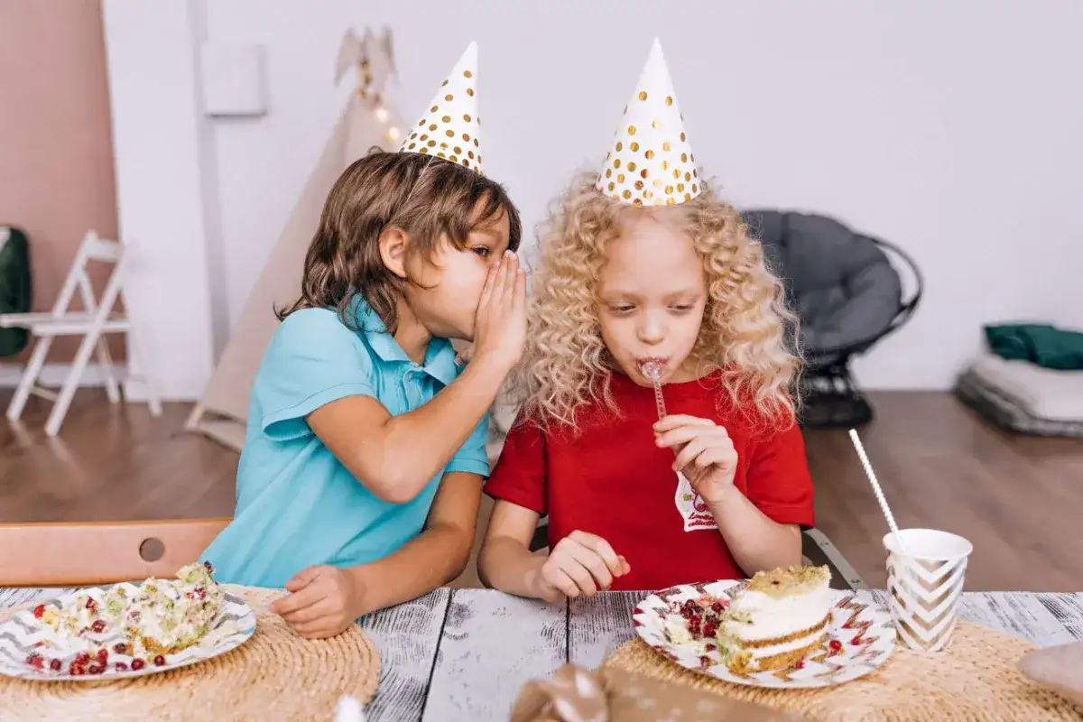 Chłopiec szepcze coś do ucha dziewczynce, która je tort. To zaproszenie na urodziny, pełne słodkich sekretów i zabawy.